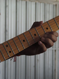 A hand is gripping the neck of a guitar, pressing down on the strings. The guitar has a wooden neck with metal frets and dot markers. The background is a corrugated metal surface, creating a contrast with the wooden texture of the guitar.