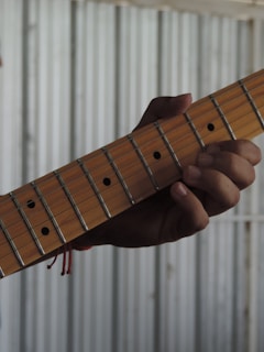 A hand is gripping the neck of a guitar, pressing down on the strings. The guitar has a wooden neck with metal frets and dot markers. The background is a corrugated metal surface, creating a contrast with the wooden texture of the guitar.