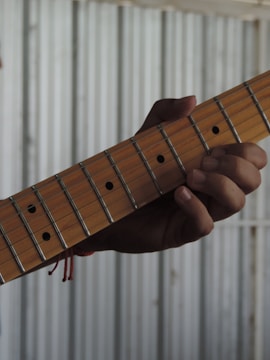 A hand is gripping the neck of a guitar, pressing down on the strings. The guitar has a wooden neck with metal frets and dot markers. The background is a corrugated metal surface, creating a contrast with the wooden texture of the guitar.