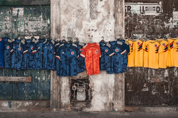 A row of football jerseys are hanging on a line against a weathered concrete wall. The jerseys are primarily blue and yellow in color with one standout red jersey in the middle. The background features an old, worn door with various stickers and signs on it.
