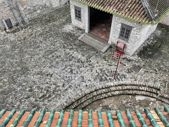 A cobblestone courtyard featuring geometric patterns is depicted. In the center-right, there is a building with white brick walls and a tiled green roof with a red and white signpost beside the entrance. The entrance is slightly elevated with steps leading to it. A ladder leans against the building on the left, and the image appears to be taken from an elevated perspective, possibly from a rooftop with green and orange tiles visible in the foreground.