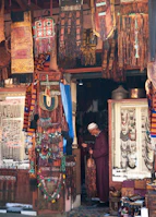A shop filled with colorful textiles, jewelry, and crafts. A person in traditional clothing stands inside, examining an item. The walls and displays are adorned with intricately designed fabrics, beads, and ornaments.