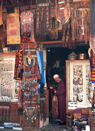 A shop filled with colorful textiles, jewelry, and crafts. A person in traditional clothing stands inside, examining an item. The walls and displays are adorned with intricately designed fabrics, beads, and ornaments.