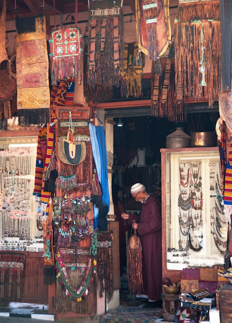 A shop filled with colorful textiles, jewelry, and crafts. A person in traditional clothing stands inside, examining an item. The walls and displays are adorned with intricately designed fabrics, beads, and ornaments.