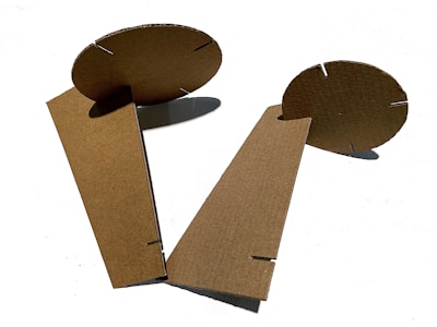 Cardboard pieces with interlocking slits form an abstract geometric structure. The material has a raw, unbleached texture, and the pieces are positioned against a white background, casting slight shadows.