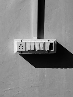 A black and white image of a wall-mounted electrical switchboard with four switches and a socket. The board is positioned against a smooth concrete wall, and there is a prominent shadow cast to the right.