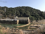 A hydroelectric dam sits against the backdrop of a densely forested mountain. The structure is built across a river with visible turbines. The surrounding area includes rocky and dry terrain, while the sky is clear.