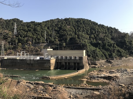 A hydroelectric dam sits against the backdrop of a densely forested mountain. The structure is built across a river with visible turbines. The surrounding area includes rocky and dry terrain, while the sky is clear.