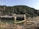A hydroelectric dam sits against the backdrop of a densely forested mountain. The structure is built across a river with visible turbines. The surrounding area includes rocky and dry terrain, while the sky is clear.