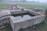 A rectangular water storage tank made of concrete is situated outdoors. Nearby, a brick structure with a metal pipe is likely serving as a water source. The area is surrounded by grass and dirt, with a slightly foggy or misty background.