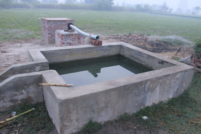 A rectangular water storage tank made of concrete is situated outdoors. Nearby, a brick structure with a metal pipe is likely serving as a water source. The area is surrounded by grass and dirt, with a slightly foggy or misty background.