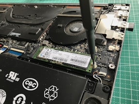 A close-up view of a laptop's internal components being worked on with a screwdriver. The image shows a solid-state drive being secured onto the motherboard. Various components of the laptop, including the cooling fan and connectors, are visible.