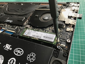 A close-up view of a laptop's internal components being worked on with a screwdriver. The image shows a solid-state drive being secured onto the motherboard. Various components of the laptop, including the cooling fan and connectors, are visible.