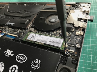 A close-up view of a laptop's internal components being worked on with a screwdriver. The image shows a solid-state drive being secured onto the motherboard. Various components of the laptop, including the cooling fan and connectors, are visible.