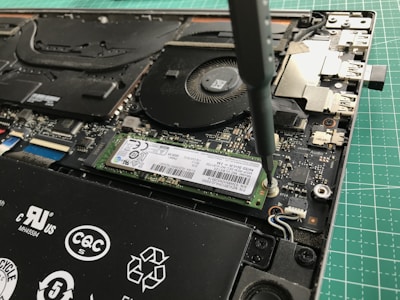 A close-up view of a laptop's internal components being worked on with a screwdriver. The image shows a solid-state drive being secured onto the motherboard. Various components of the laptop, including the cooling fan and connectors, are visible.