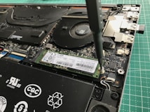 A close-up view of a laptop's internal components being worked on with a screwdriver. The image shows a solid-state drive being secured onto the motherboard. Various components of the laptop, including the cooling fan and connectors, are visible.