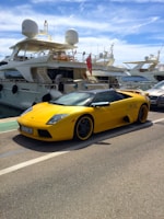 A shiny sports car parked beside a marina with yachts in the background.