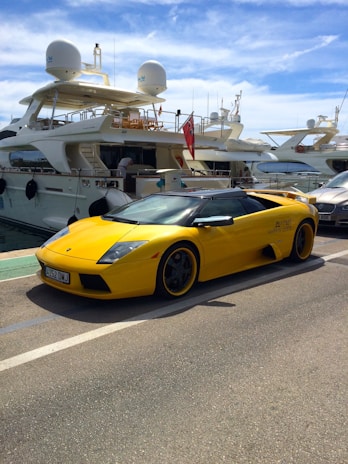 A bright yellow luxury sports car is parked on a marina, adjacent to several large yachts docked alongside a pier. The sports car features sleek lines and a low profile. The sky is partly cloudy, adding a serene backdrop to the scene. The marina setting suggests a location of wealth and leisure.
