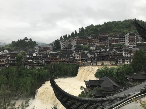 A scenic view of a town built on a hillside, with the buildings densely packed together. In the foreground, a wide, powerful waterfall cascades down, flanked by lush greenery. Traditional architectural elements such as pagodas and curved roofs are visible atop the buildings. The landscape is lush and green, with a thick forest covering the hill behind the town. The sky is overcast, adding a moody atmosphere to the image.