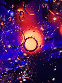 A vibrant abstract image featuring large and small bubbles suspended in a liquid. The bubbles are illuminated by an intense blend of blue, purple, and orange light, creating a striking contrast. Tiny droplets are scattered across the scene, enhancing the dynamic and somewhat otherworldly appearance.