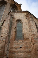 A tall, detailed brick wall of a historical building featuring arched windows with diamond-patterned glass panes. The architecture includes Gothic elements with intricate masonry and a prominent roofline.