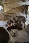 An ornate pipe organ is mounted on the stone wall of a cathedral, with intricate woodwork and decorative carvings. The arched ceiling and large stained-glass window allow natural light to illuminate the interior.