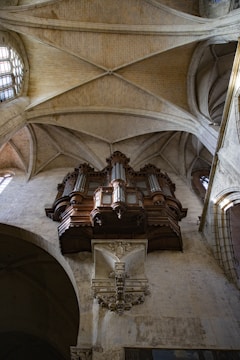 An ornate pipe organ is mounted on the stone wall of a cathedral, with intricate woodwork and decorative carvings. The arched ceiling and large stained-glass window allow natural light to illuminate the interior.