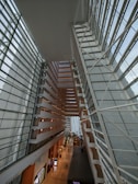 An expansive, modern architectural interior with geometric lines and large glass surfaces. The structure features multiple levels, with angular designs and a vast open space allowing natural light to flood the area. People walk along a corridor on a lower level, giving the impression of a bustling environment.