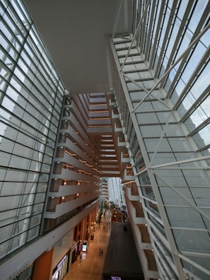 An expansive, modern architectural interior with geometric lines and large glass surfaces. The structure features multiple levels, with angular designs and a vast open space allowing natural light to flood the area. People walk along a corridor on a lower level, giving the impression of a bustling environment.