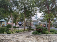 A landscaped area with lush greenery, including trees and bushes, surrounds a paved stone pathway. In the background, a large spherical sculpture with letters partially visible is situated near modern architectural structures characterized by tall, peaked roofs and metal elements.