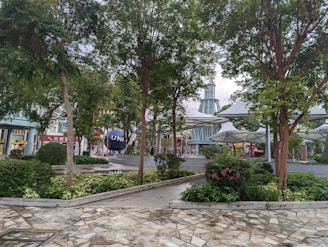 A landscaped area with lush greenery, including trees and bushes, surrounds a paved stone pathway. In the background, a large spherical sculpture with letters partially visible is situated near modern architectural structures characterized by tall, peaked roofs and metal elements.