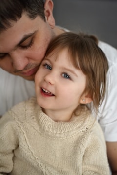 A young child with light brown hair and blue eyes is smiling while being held closely by an adult. The child is wearing a cozy, cream-colored sweater and appears joyful. The adult's face is partially visible, with a gentle expression.