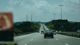 A highway stretches into the distance with multiple lanes of vehicles. There are cars traveling in both directions, and the road is bordered by lush green hills. Overhead, power lines supported by tall pylons create a backdrop against a partly cloudy sky. Streetlights line both sides of the road.