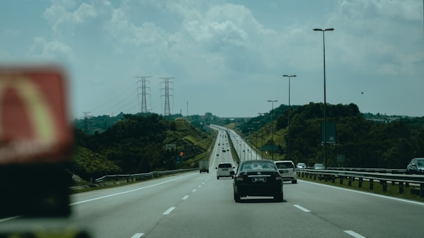A highway stretches into the distance with multiple lanes of vehicles. There are cars traveling in both directions, and the road is bordered by lush green hills. Overhead, power lines supported by tall pylons create a backdrop against a partly cloudy sky. Streetlights line both sides of the road.