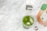 A glass with a vibrant green beverage and ice cubes is set on a marble surface. Next to it, there is a carton labeled 'Boxed Water Is Better' and a colorful matcha tea package marked 'Matcha Fresca'. A silver measuring spoon rests nearby.