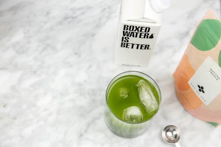 A glass with a vibrant green beverage and ice cubes is set on a marble surface. Next to it, there is a carton labeled 'Boxed Water Is Better' and a colorful matcha tea package marked 'Matcha Fresca'. A silver measuring spoon rests nearby.