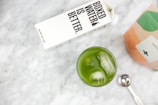 A carton of Boxed Water next to a glass filled with green matcha tea and ice cubes on a marble surface. There is also a metallic measuring spoon and a partially visible bag of matcha powder.