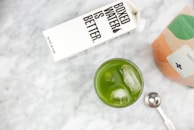 A carton of Boxed Water next to a glass filled with green matcha tea and ice cubes on a marble surface. There is also a metallic measuring spoon and a partially visible bag of matcha powder.
