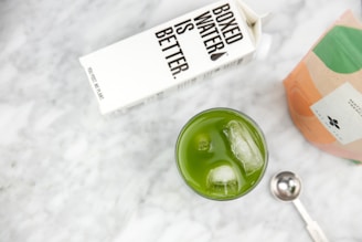 A carton of Boxed Water next to a glass filled with green matcha tea and ice cubes on a marble surface. There is also a metallic measuring spoon and a partially visible bag of matcha powder.