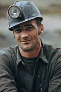 A man wearing a miner's helmet with a headlamp, dressed in a work shirt, and smiling slightly. He appears to be outdoors or in a rugged environment.