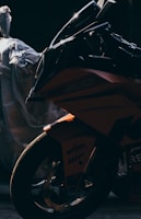 A motorcycle is captured in low lighting, emphasizing the sleek and sharp design of its fairing and front wheel. The motorcycle is partially obscured by shadows, creating a sense of mystery. A plastic-wrapped object sits in the background, adding to the contrast between the motorcycle’s glossy finish and the muted surroundings.