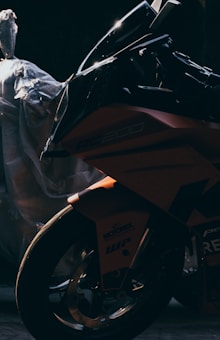 A motorcycle is captured in low lighting, emphasizing the sleek and sharp design of its fairing and front wheel. The motorcycle is partially obscured by shadows, creating a sense of mystery. A plastic-wrapped object sits in the background, adding to the contrast between the motorcycle’s glossy finish and the muted surroundings.