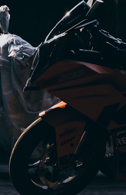 A motorcycle is captured in low lighting, emphasizing the sleek and sharp design of its fairing and front wheel. The motorcycle is partially obscured by shadows, creating a sense of mystery. A plastic-wrapped object sits in the background, adding to the contrast between the motorcycle’s glossy finish and the muted surroundings.