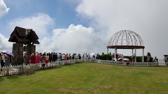A group of people is gathered in an open grassy area surrounded by a white picket fence. There are two structures: one resembling a small stone building with a pitched roof on the left, and an open dome-like gazebo on the right.