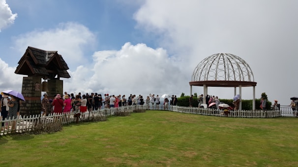 A group of people is gathered in an open grassy area surrounded by a white picket fence. There are two structures: one resembling a small stone building with a pitched roof on the left, and an open dome-like gazebo on the right.