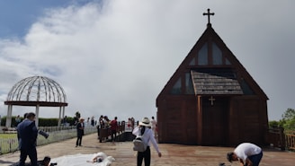 A small wooden chapel with a cross on top is situated on a wooden deck. People are gathered around the chapel and a white metal gazebo structure can be seen in the background. Some individuals appear to be setting up for a photoshoot, with a person lying on the ground in a white outfit.