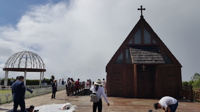 A small wooden chapel with a cross on top is situated on a wooden deck. People are gathered around the chapel and a white metal gazebo structure can be seen in the background. Some individuals appear to be setting up for a photoshoot, with a person lying on the ground in a white outfit.