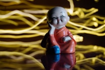 A small statue of a joyful, smiling monk in a traditional robe is seated with one hand resting on his cheek. The background features dynamic, golden light streaks that create an energetic and lively atmosphere. The statue stands on a glossy surface, reflecting parts of its image.