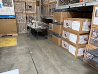 A warehouse setting with stacks of items in cardboard boxes. Some boxes contain outdoor furniture like chairs. There are also large industrial fans or air conditioning units wrapped in plastic on the left side. The floor is concrete, and there's a marked yellow area.