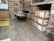 A warehouse setting with stacks of items in cardboard boxes. Some boxes contain outdoor furniture like chairs. There are also large industrial fans or air conditioning units wrapped in plastic on the left side. The floor is concrete, and there's a marked yellow area.
